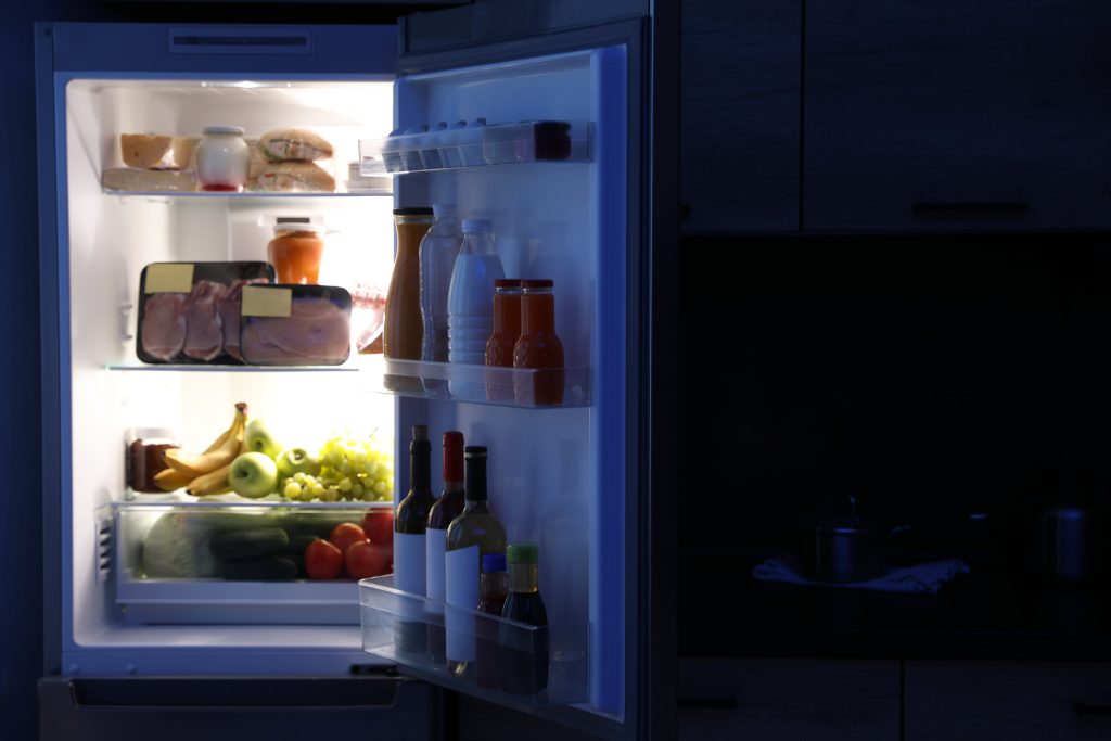 A refrigerator standing open in a dark kitchen, its interior light showing shelves of food and bottles in the door
