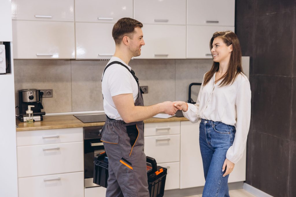A tradesman in overalls shaking hands with a homeowner in a kitchen, tool case at his feet.