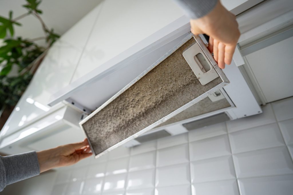 Two hands lifting a grease-caked mesh filter out of a slide-out cooker hood.