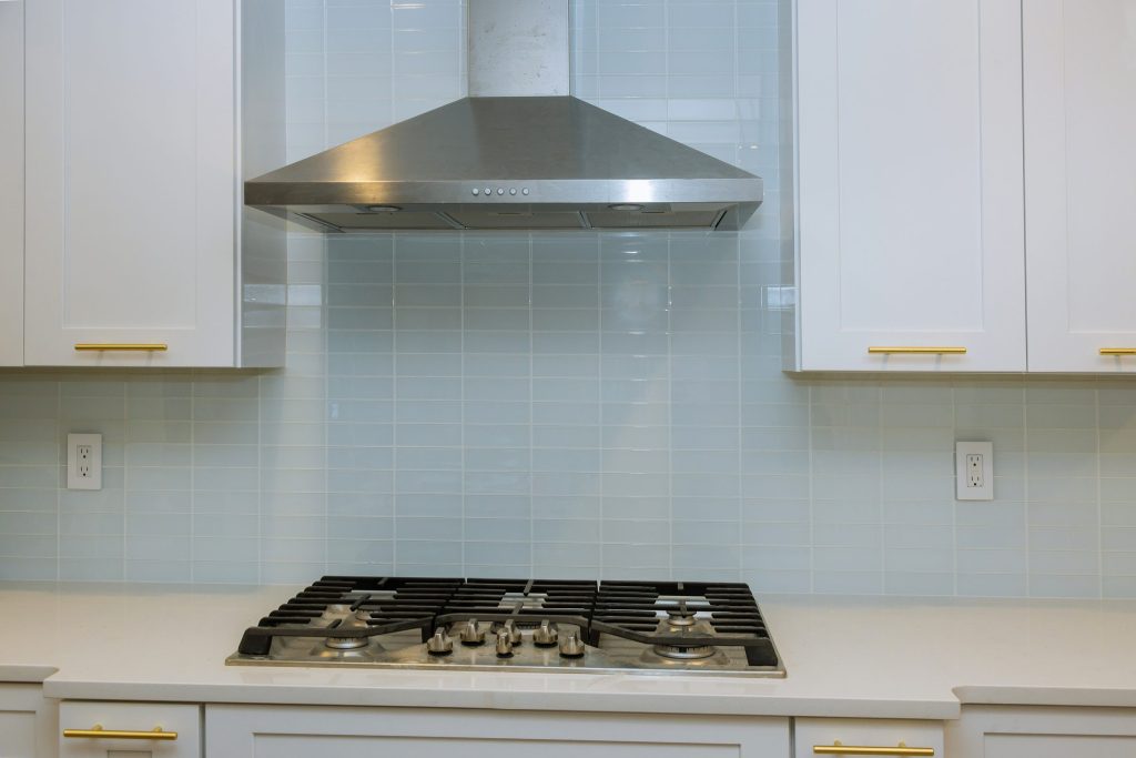 A stainless hood mounted over a gas cooktop in a white tiled kitchen