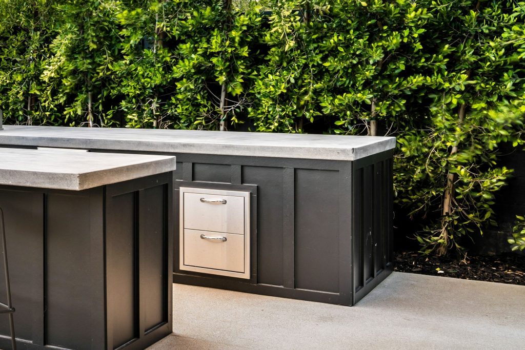 A stainless two-drawer unit built into a dark outdoor kitchen island against a hedge