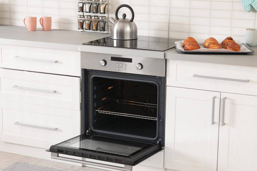 A stainless oven open in a white kitchen with a tray cooling above it