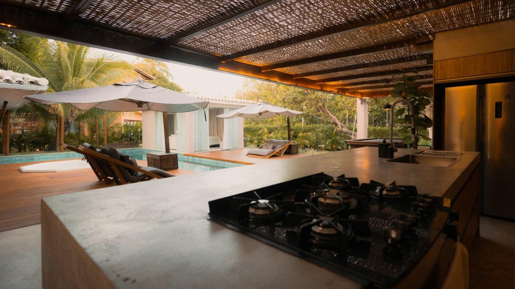 Covered outdoor kitchen beside a pool, with a built-in cooktop on a concrete counter