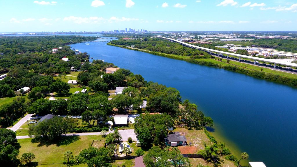 A wide waterway between wooded houses with a city skyline far off, from the air