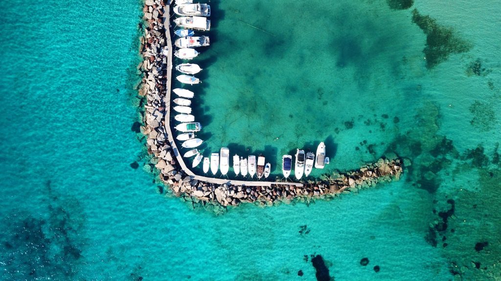Boats moored inside a rock breakwater over clear green shallows, straight down