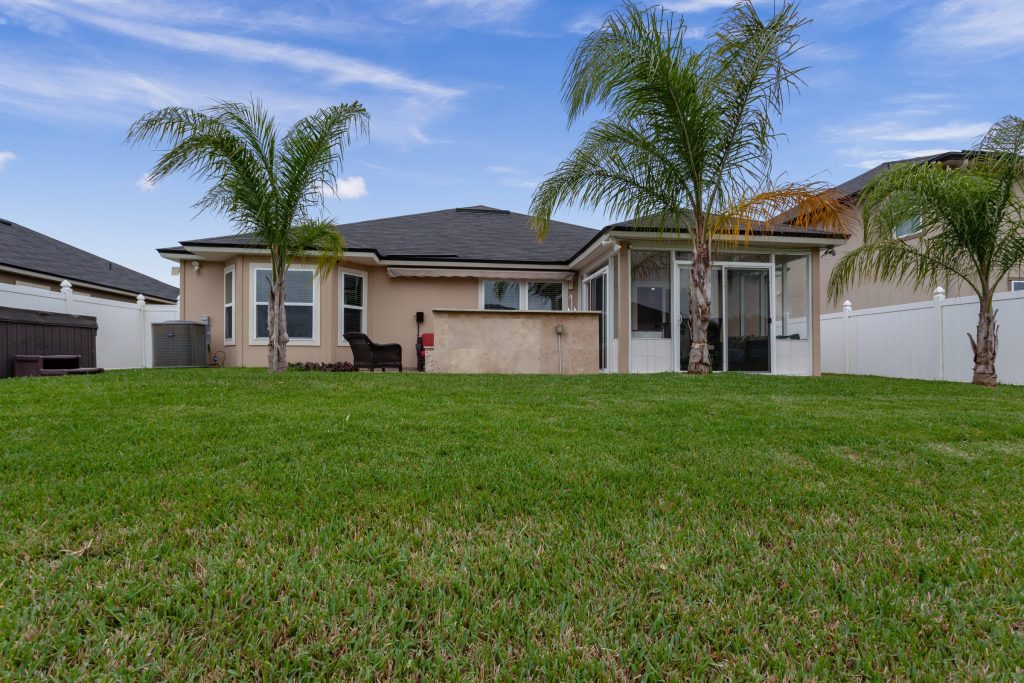 A single-story house behind a wide lawn with palms and a white fence