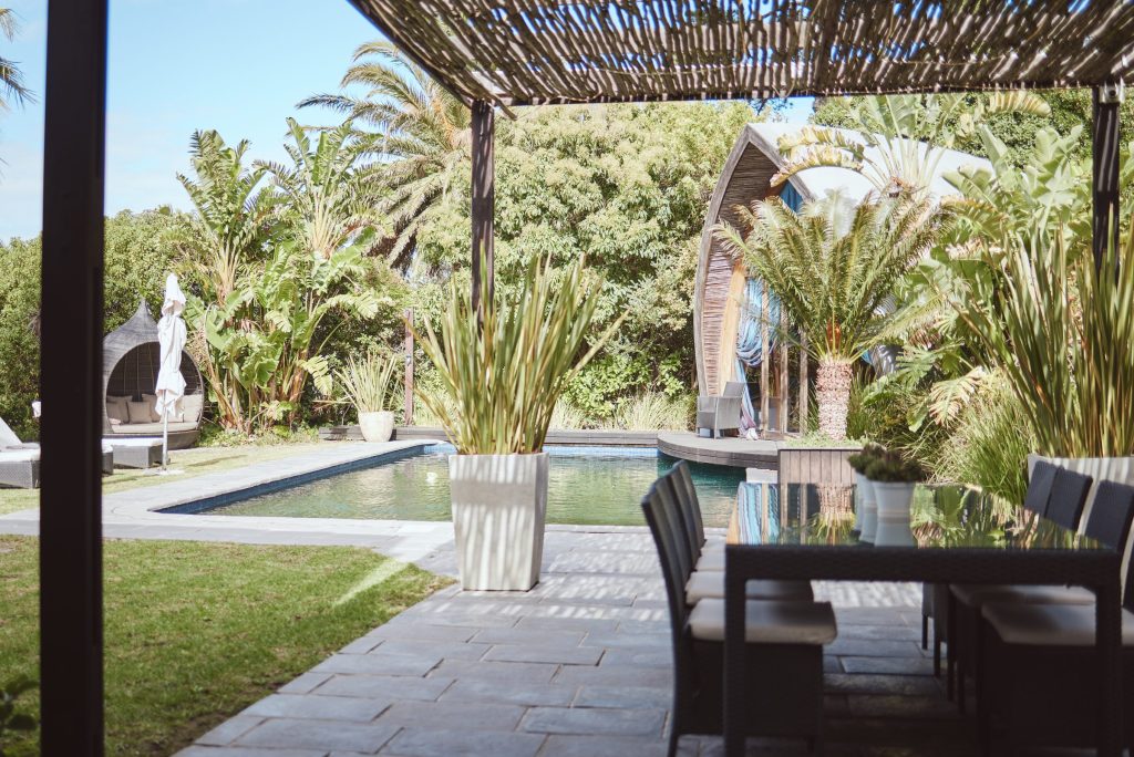 A shaded pergola and dining table beside a pool in a planted garden