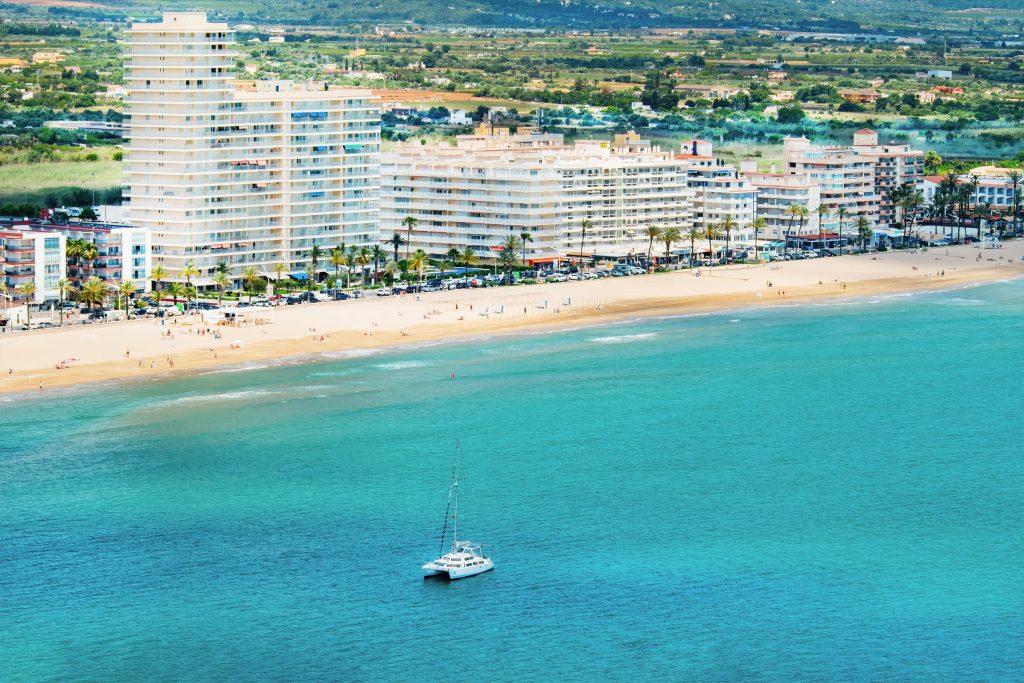 A long curve of beach with apartment blocks behind it and a catamaran offshore