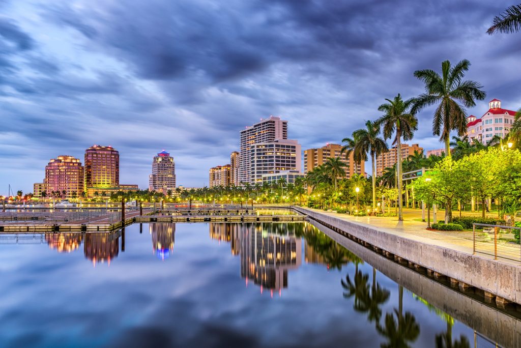 A lit waterfront skyline reflected in still water at dusk, palms along the walk