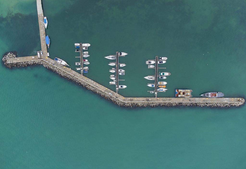 Yachts along two arms of a breakwater in a shallow basin, from above