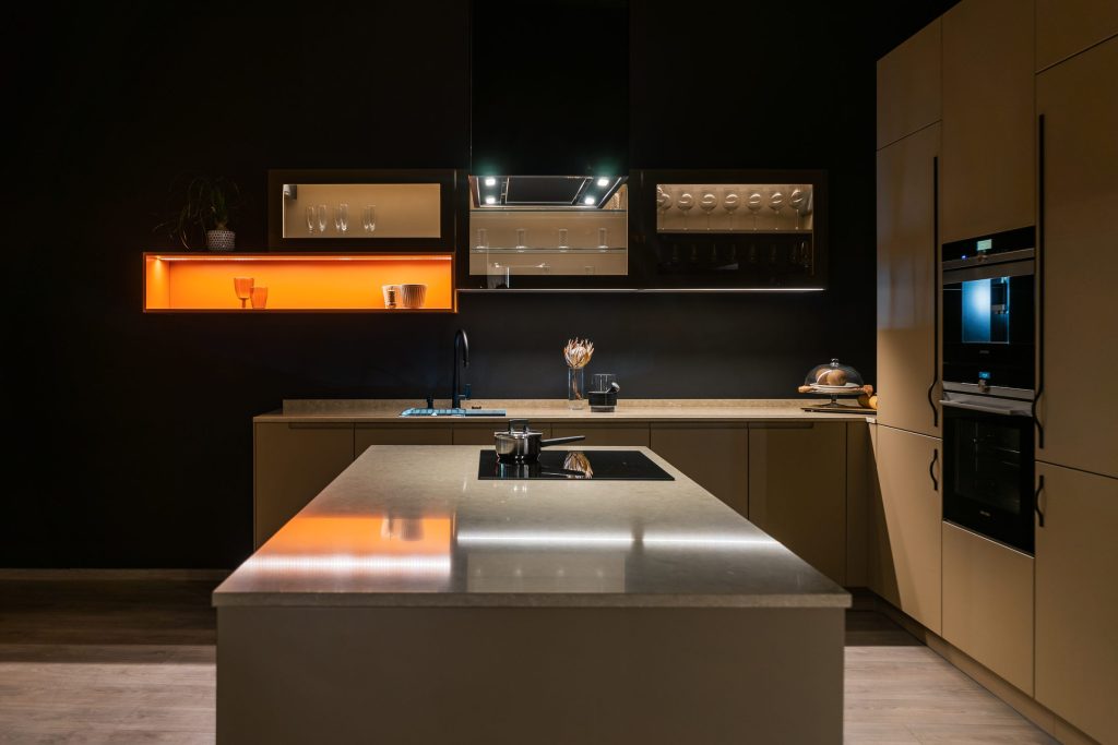 A dark contemporary kitchen lit by warm strip lighting, steel island, tall oven column.