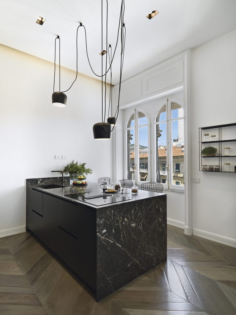 A white room with a black marble island, three black pendants on long flex, arched windows onto a town.