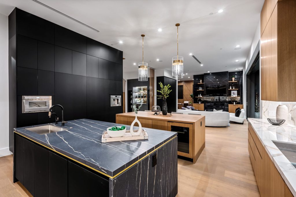 A black kitchen with a marble island edged in brass, a steam oven and a coffee machine built into the wall, opening onto a living room.
