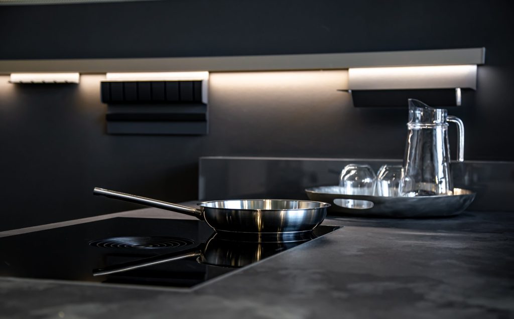A steel pan on a black hob in a dark kitchen, a lit strip along the wall behind it