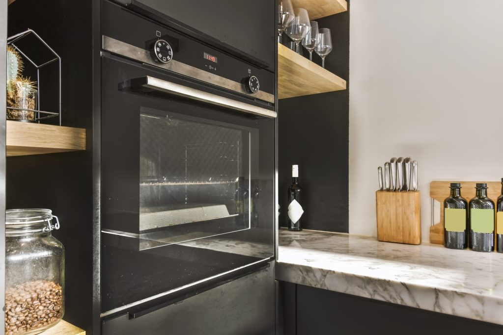A dark kitchen with a built-in oven in black cabinetry, oak shelves and a marble counter beside it