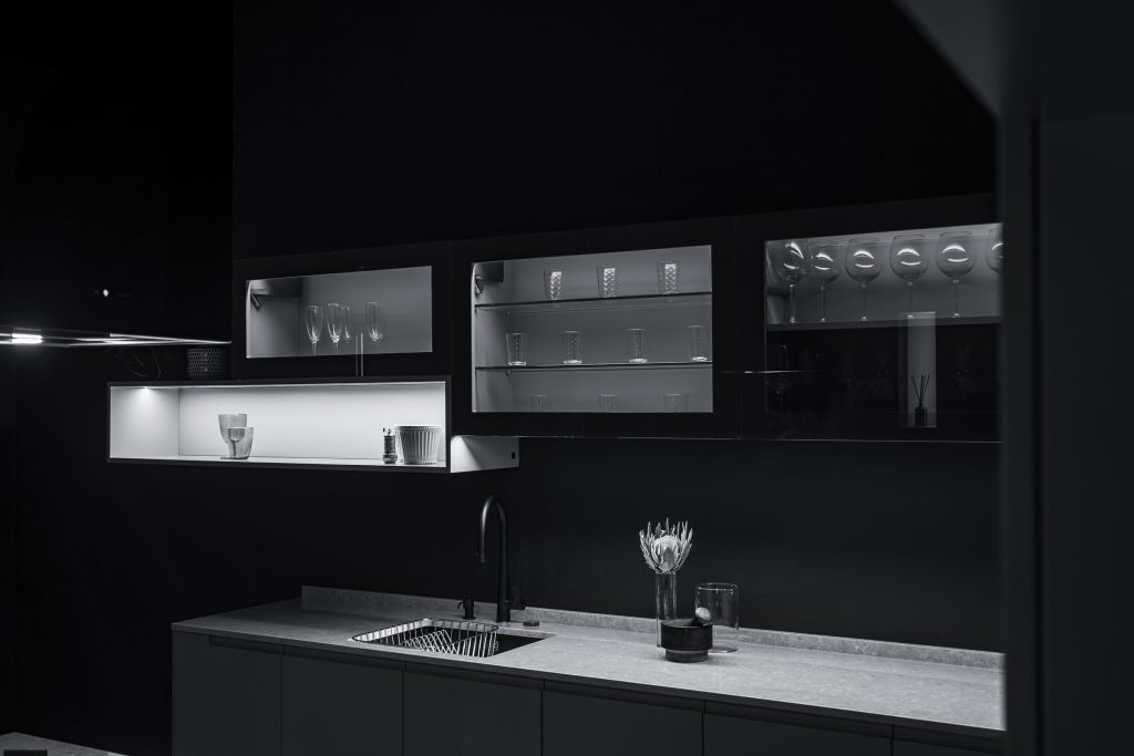 A dimly lit minimalist kitchen, a stone worktop and lit glass-fronted wall units.
