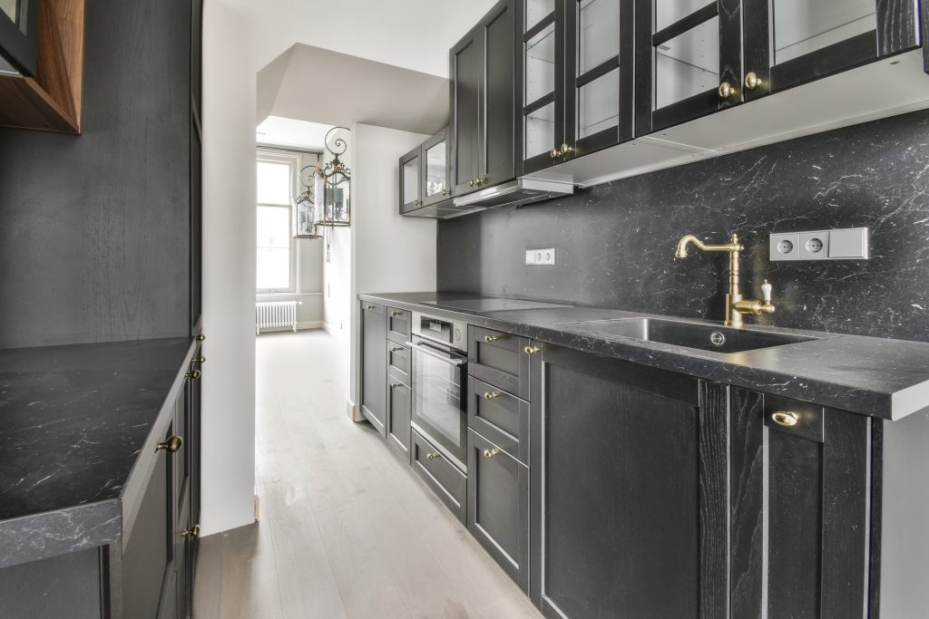 A narrow black kitchen with brass knobs and a black marble worktop and splashback, an oven under the induction hob.