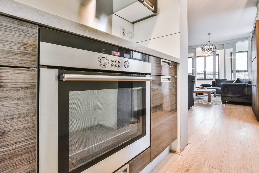 A stainless wall oven in wood cabinetry, living room beyond.