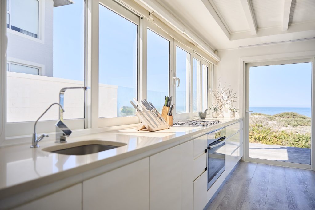 A white galley kitchen along a run of windows onto the sea, a gas hob and an oven in the counter.