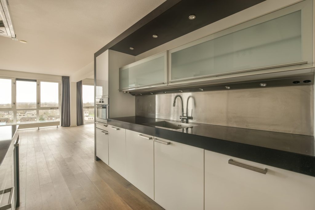 A white kitchen with a stainless splashback and glass wall cabinets, black worktop, city windows behind.