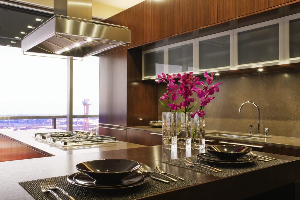 A walnut kitchen at night with a brass island hood over a gas hob, orchids on the counter and a city window.