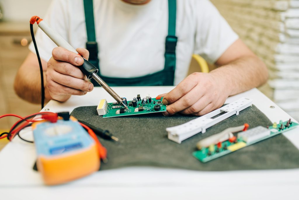 A technician soldering a green control board at a bench, a multimeter and spare boards beside it