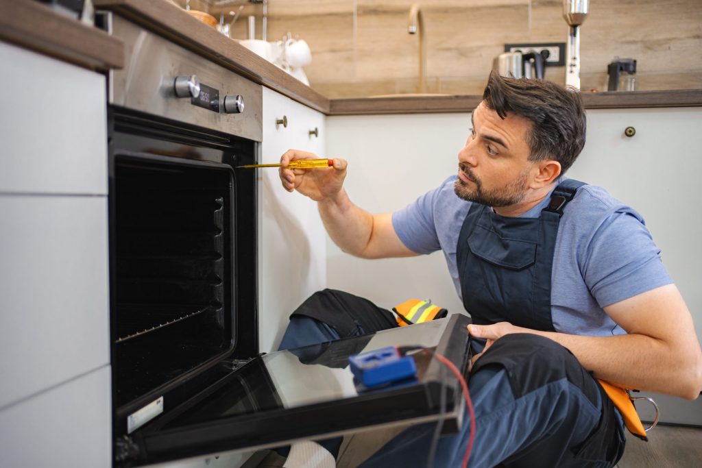 A technician kneeling at an open built-in oven in a home kitchen, screwdriver in hand and a tool roll on the floor