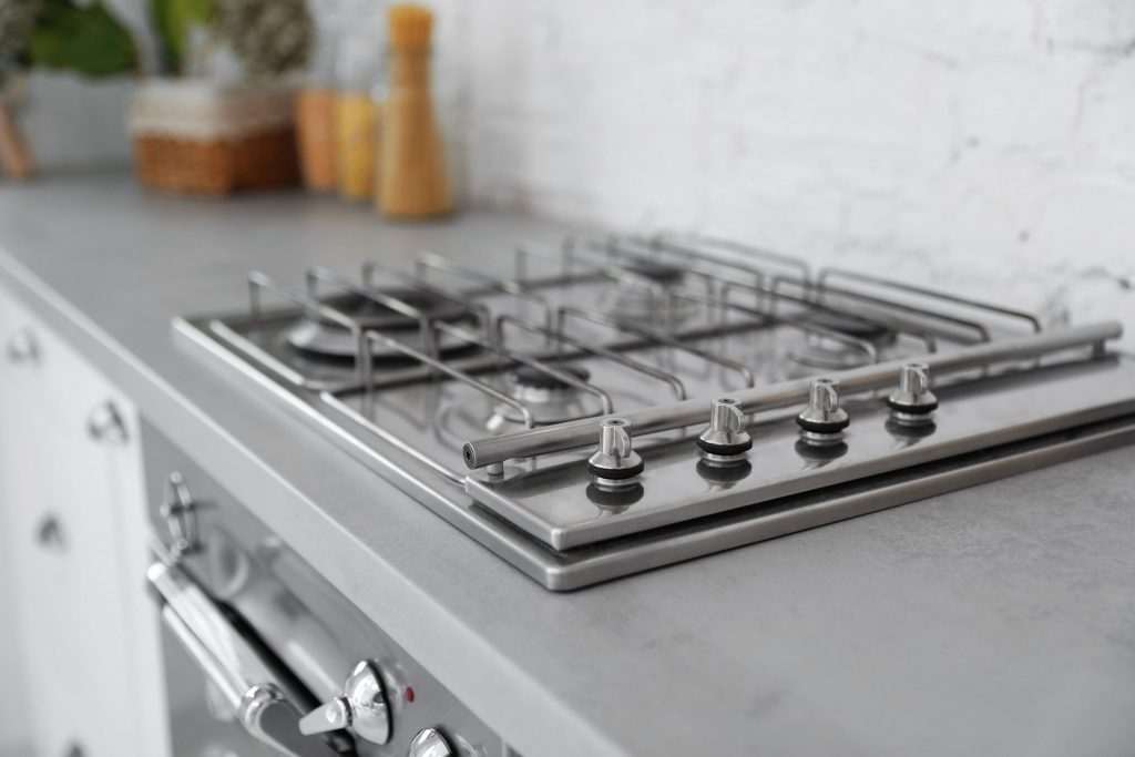 A stainless gas cooktop with four burners set into a grey worktop, brick wall behind, a range front below.
