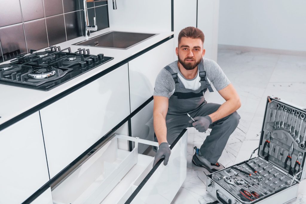 A fitter kneeling at a base unit below a gas hob, tool case open on the floor beside him