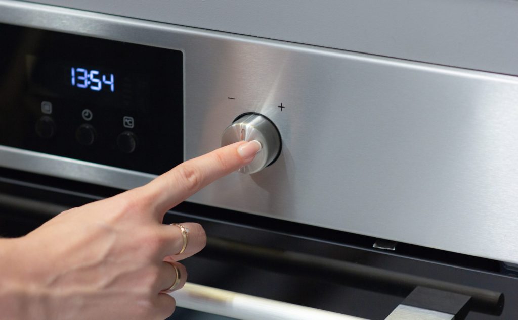 A finger on the round control of a stainless wall oven, the display reading 13:54.