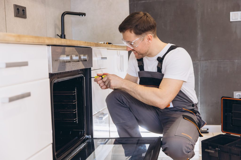 A fitter in safety glasses kneeling at a built-in wall oven, driving a screw into the cabinet frame.