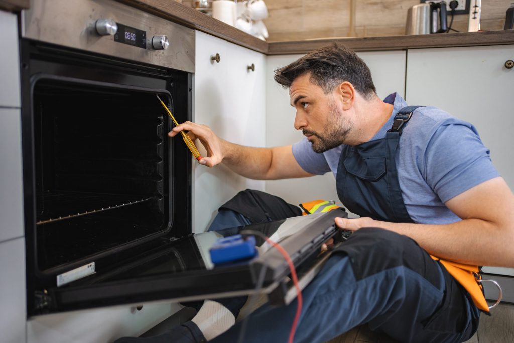 A technician kneeling at an open built-in oven with a multimeter, probes on the cavity wiring