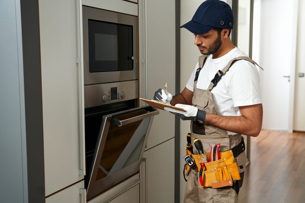 A technician with a clipboard beside an open built-in oven and microwave column, tool belt on