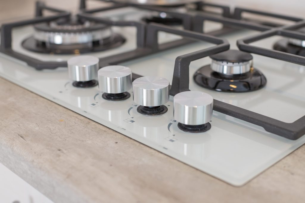 A white glass gas hob with cast grates and four brushed steel knobs, close up on the front edge.