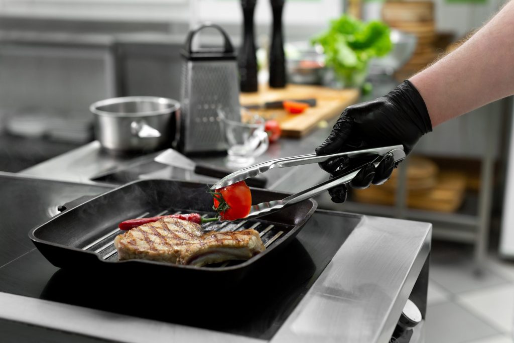 A gloved hand turning food with tongs on a ribbed griddle plate set into a cooktop