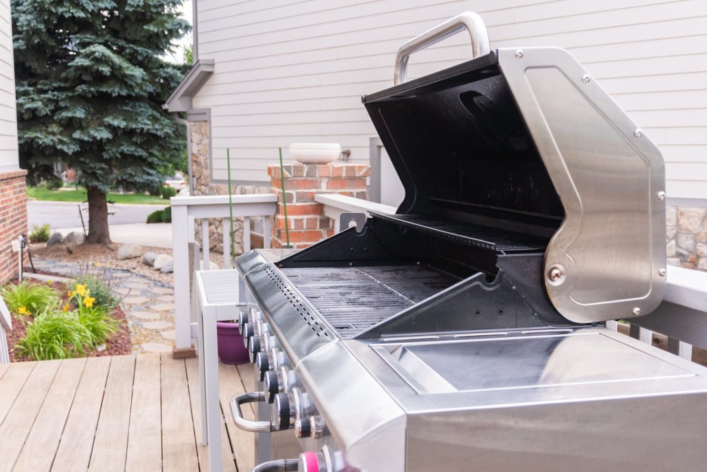 A stainless outdoor gas grill on a deck with the lid raised, burner tubes and control knobs in view