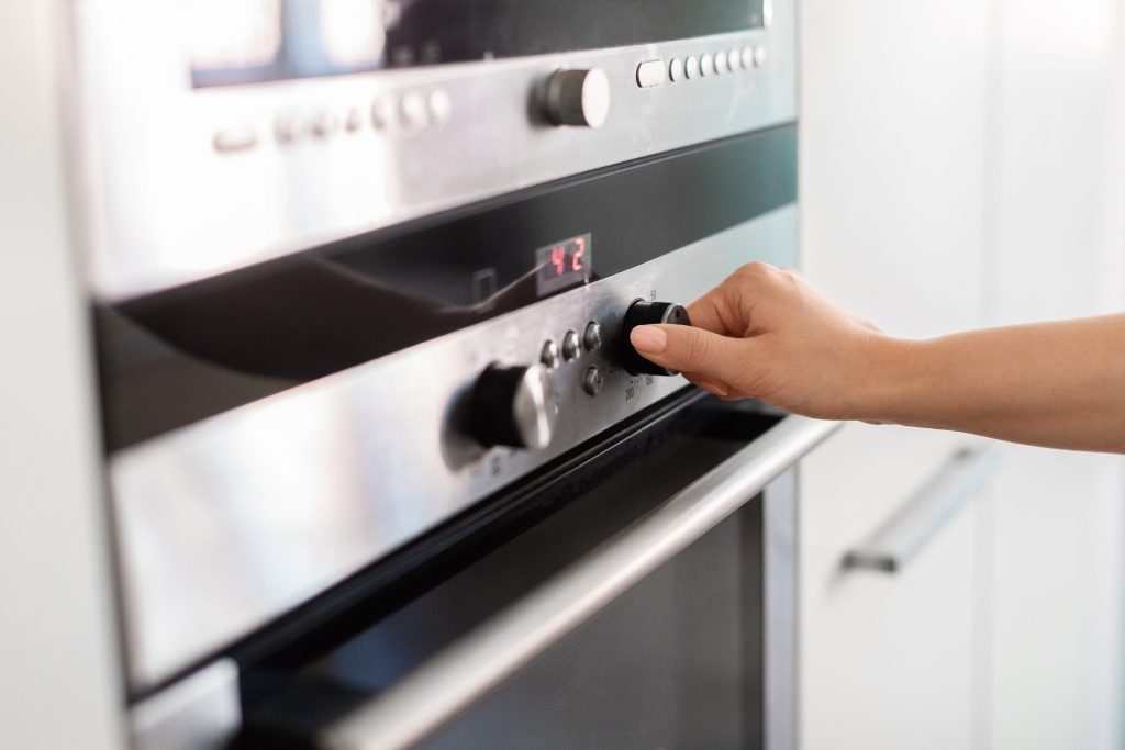 A hand turning the temperature dial on a stainless wall oven, red display lit.