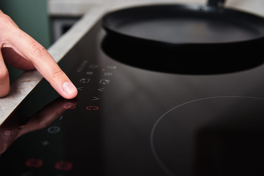 A finger on the slider of an induction hob, red digits lit, a pan out of focus behind.