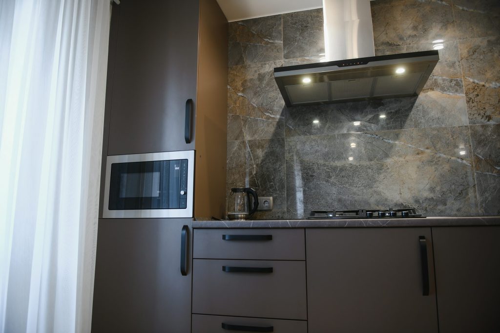 An angled stainless hood lit over a gas hob against a stone splashback in a dark kitchen